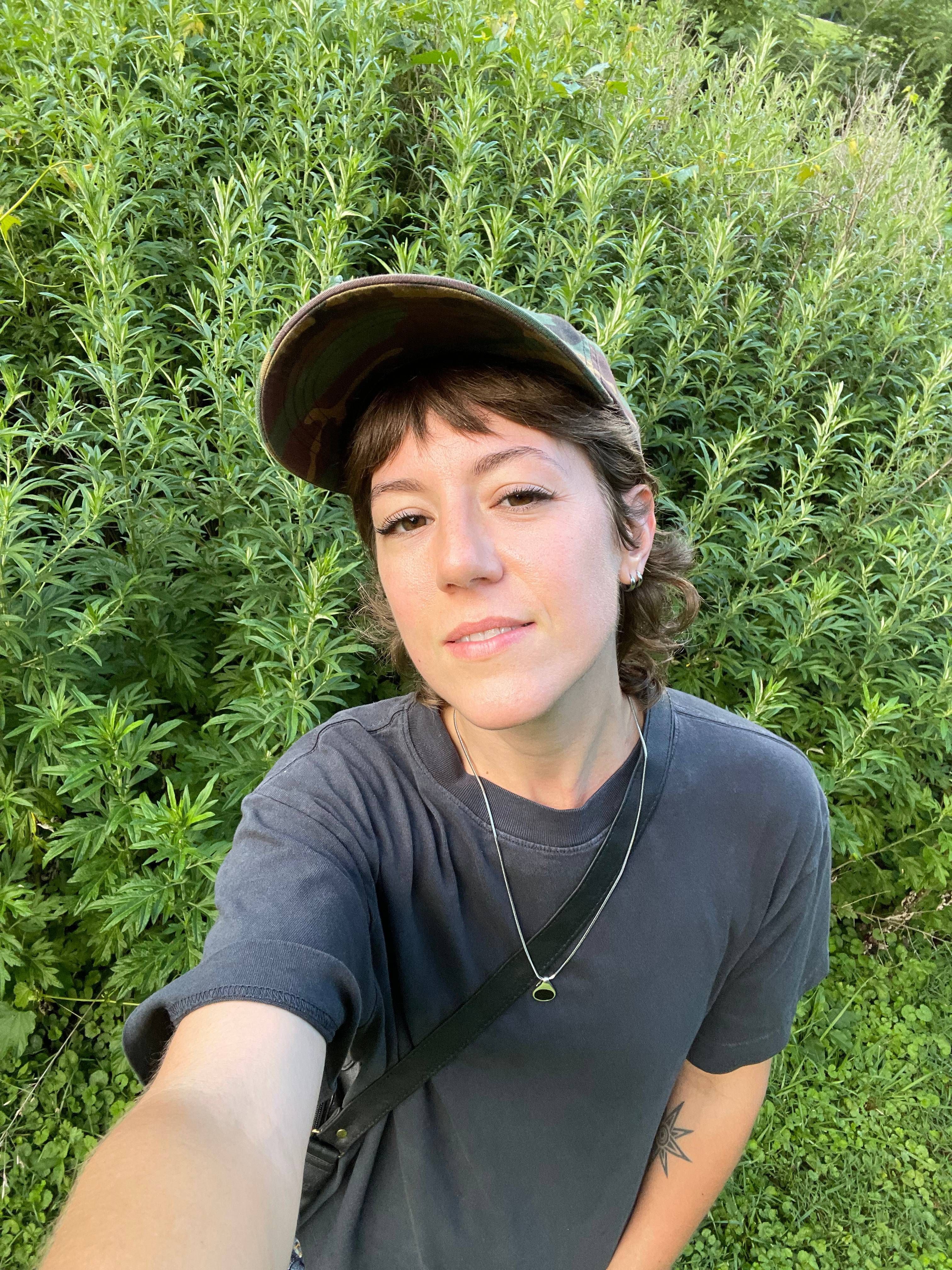 a selfie photo of Leah standing in front of a field of wild mugwort. they're wearing a grey t-shirt and camo baseball hat